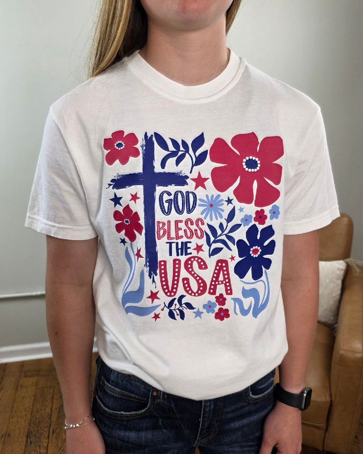 The God Bless the USA Graphic Tee by Cosmos + Clover Boutique is a white Comfort Colors youth shirt with a cross, patriotic red, white, and blue flowers, and stars—perfect for a classic Americana look.