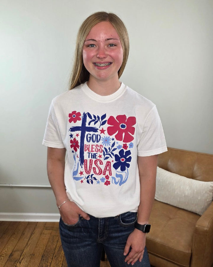 A teen girl with blonde hair smiles indoors near a tan couch, wearing the Cosmos + Clover Boutique God Bless the USA Graphic Tee featuring red, blue, and pink floral designs.