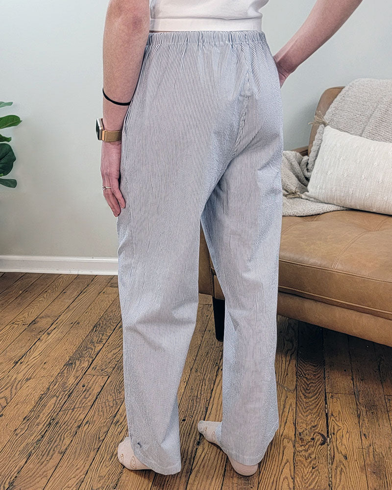 A person stands on a wooden floor near a tan sofa, wearing the Love Poem Vedea Pinstripe Pant. Only their lower back and legs in the light blue and white striped pants are visible.
