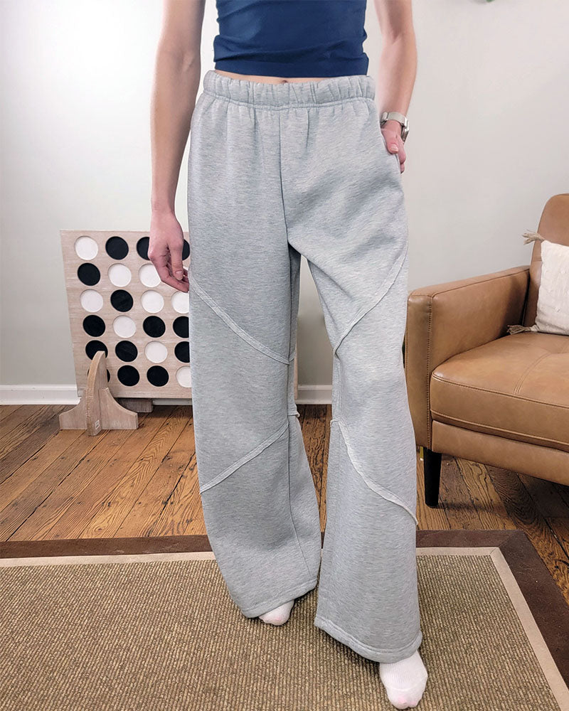 A person wearing HYFVE, Inc’s Sloan Wide Leg Pants in heather grey stands indoors on a brown rug next to a tan armchair and large Connect Four game. Only the lower torso and legs are visible.