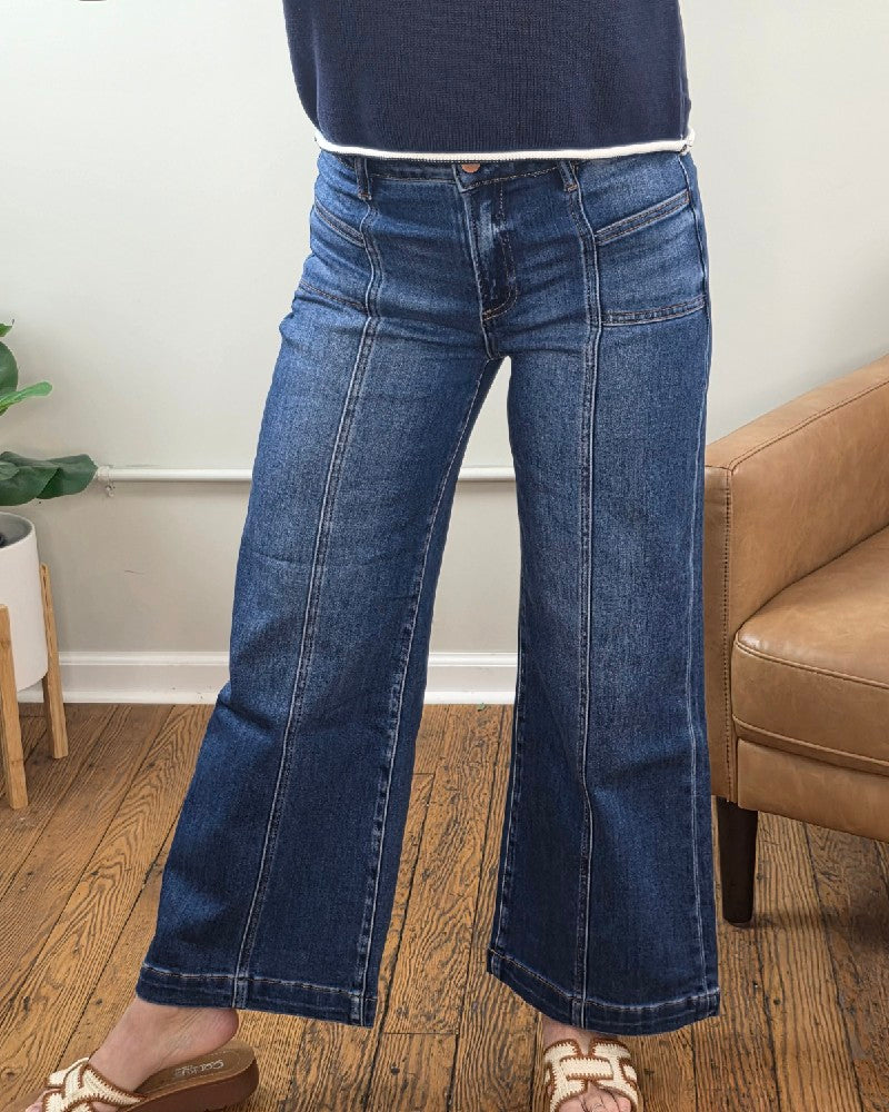 A person wearing Hallie Crop Wide Leg Jeans with Front Patch Pocket from Risen and a navy top stands indoors on a wooden floor by a tan armchair and green potted plant, with only their lower half visible.