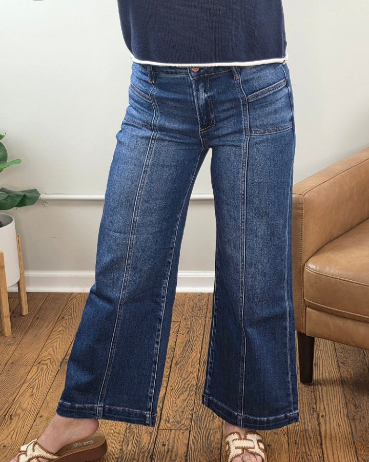 A person wearing Hallie Crop Wide Leg Jeans with Front Patch Pocket from Risen and a navy top stands indoors on a wooden floor by a tan armchair and green potted plant, with only their lower half visible.