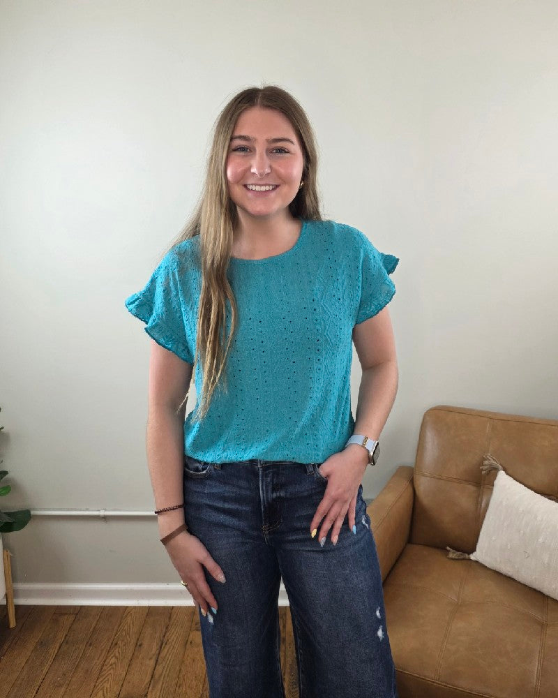 A young woman with long brown hair smiles in a living room, wearing the Lovesoft Inga Eyelet Single Ruffle Short-Sleeve Top. A tan couch with a white pillow and a plant are visible in the background.