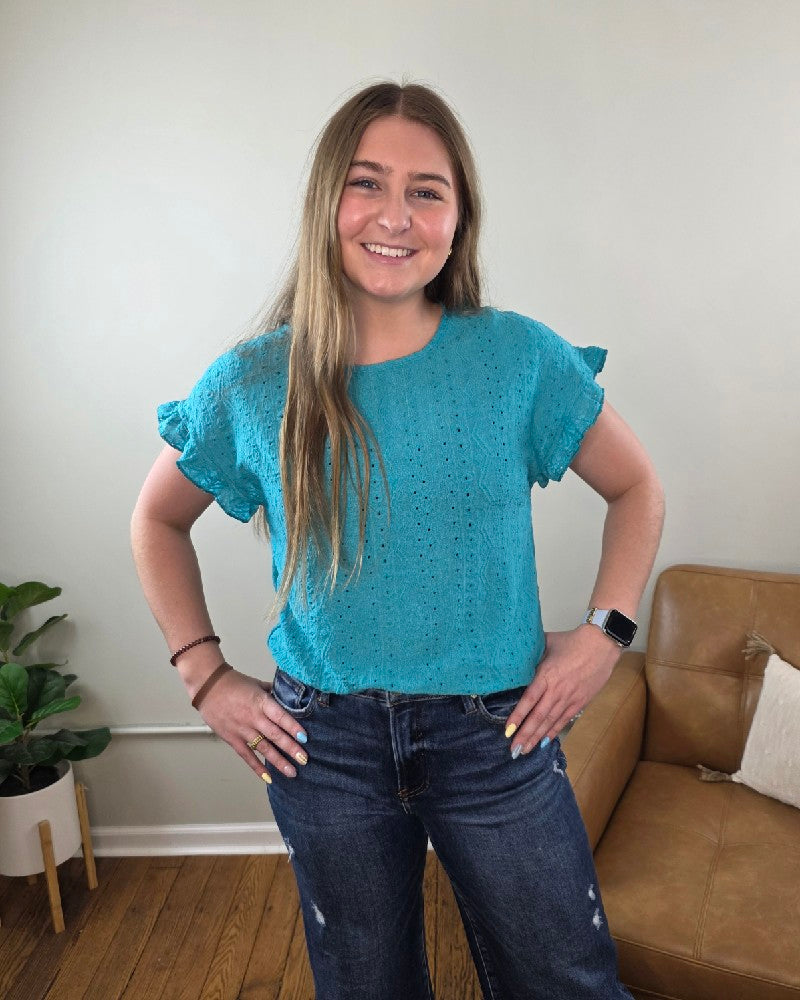 A woman with long, straight hair smiles at the camera indoors, wearing the Lovesoft Inga Eyelet Single Ruffle Short-Sleeve Top in turquoise blue, dark jeans, and a smartwatch. Behind her are a tan couch and a potted plant.
