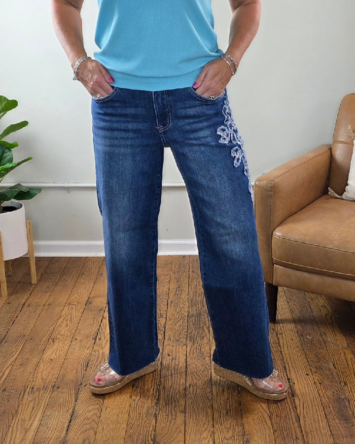 A person wearing the Iris Crop Straight Flower Patch Jeans from Risen, paired with a light blue top and wedge sandals, stands on a wooden floor next to a tan chair and a leafy plant.