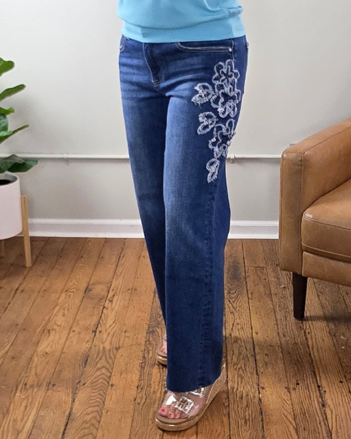 Wearing the Iris Crop Straight Flower Patch Jeans from Risen—mid-rise, blue stretch denim featuring white floral embroidery on the right thigh—paired with wedge sandals, this individual stands on a wooden floor beside a tan chair and a plant.