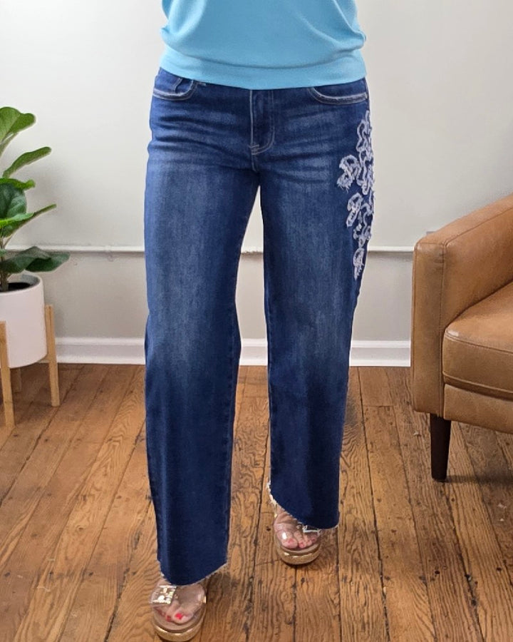 A person wearing the Iris Crop Straight Flower Patch Jeans from Risen—featuring white embroidery on the left thigh—paired with a light blue top and clear sandals, stands on a wooden floor near a tan armchair and a green potted plant.