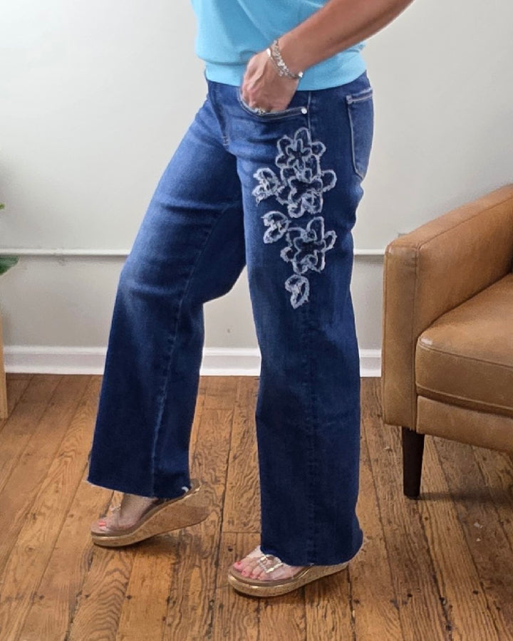 Wearing the Iris Crop Straight Flower Patch Jeans from Risen, which feature white floral embroidery on one leg, a person stands on a wooden floor by a tan chair. The stretch denim is styled with a blue top and sandals.