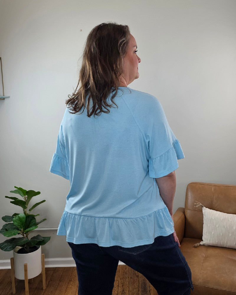 A woman with shoulder-length brown hair stands with her back to the camera in a living room, wearing the Shewin Isabella Short Sleeve Ruffle Blouse in light blue and dark jeans, surrounded by a plant and brown couch.