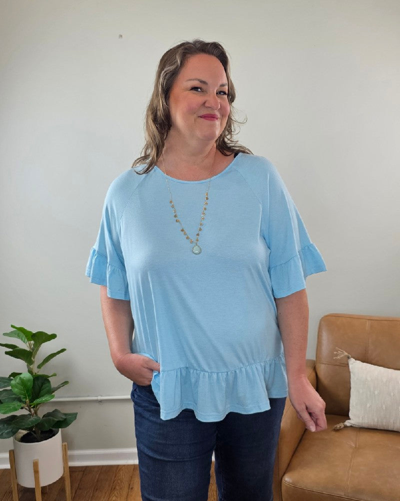 A woman in the Shewin Isabella Short Sleeve Ruffle Blouse and dark jeans stands indoors, smiling. A long gold necklace, tan chair with a white cushion, and potted plant appear in the background.