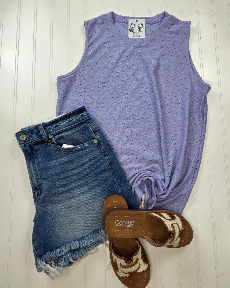 Flat lay of the Lovesoft Isla Lavender Eyelet Tank Top, featuring an eyelet design and side knot, paired with distressed blue denim shorts and brown sandals with white woven details on a white wooden background.