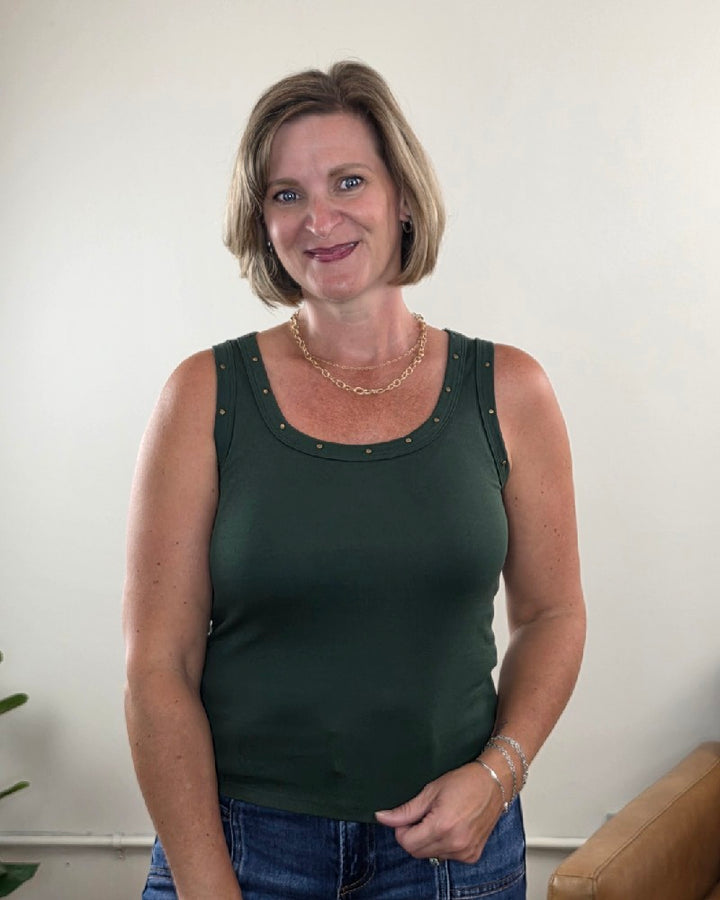 A woman with short light brown hair is standing indoors, smiling at the camera while wearing the Jessica Studded Tank Top by Timing—an olive scoop-neck tank featuring bronze studded details. She accessorizes with a gold necklace and bracelets, and a brown chair and a plant can be seen in the background.