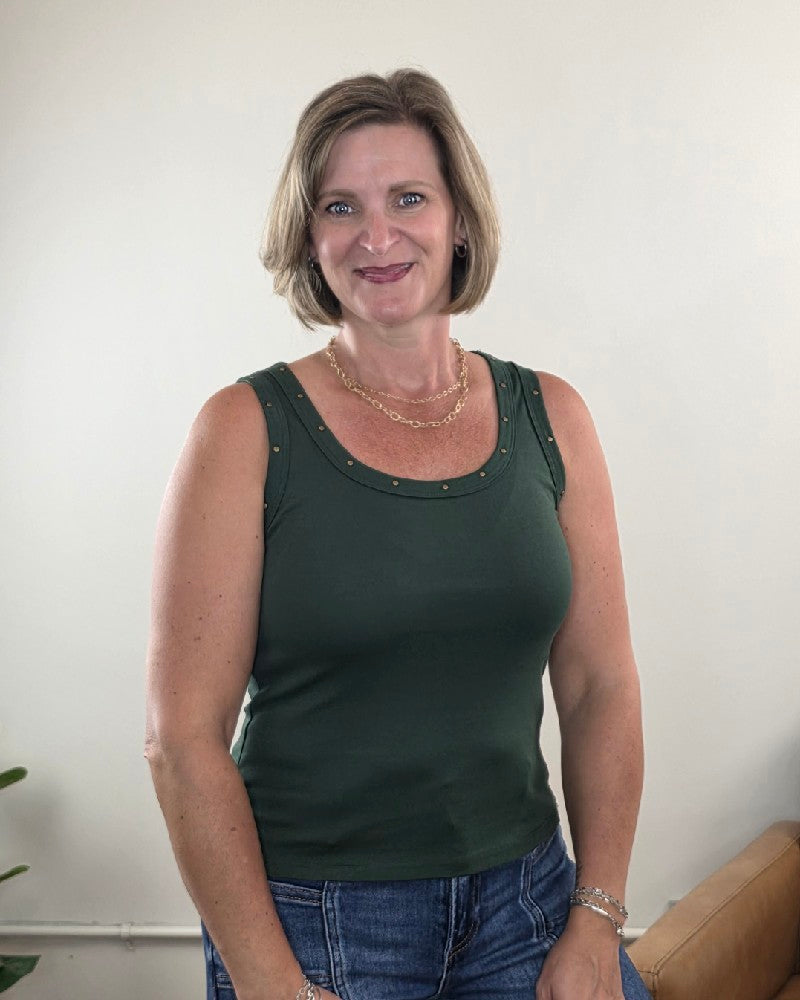A woman with short, light brown hair stands smiling in front of a plain light-colored wall, wearing the Timing Jessica Studded Tank Top—an olive, scoop-neck style—paired with blue jeans. A plant is partially visible in the bottom left corner.