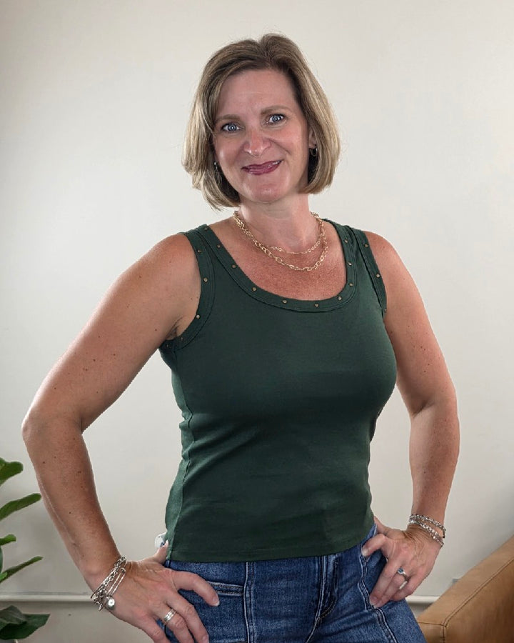 A woman with short blonde hair smiles at the camera, wearing the Jessica Studded Tank Top by Timing in olive, paired with blue jeans and jewelry, standing indoors against a light-colored wall with a plant partially visible.