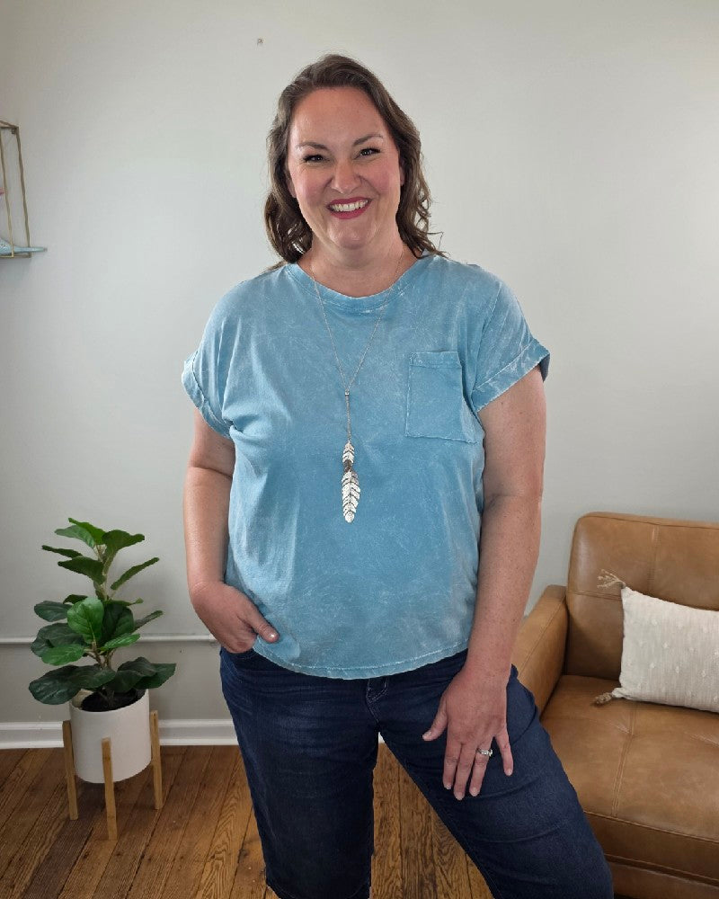 A woman with wavy brown hair smiles in a cozy room, wearing the ZENANA Jessie Crinkle Washed Rolled Short Sleeve Top in light blue, dark jeans, and a long feather necklace, with one hand in her pocket by a plant and tan chair.