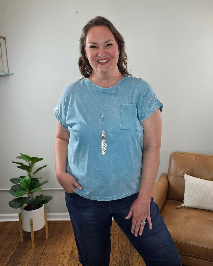A woman with wavy brown hair smiles in a cozy room, wearing the ZENANA Jessie Crinkle Washed Rolled Short Sleeve Top in light blue, dark jeans, and a long feather necklace, with one hand in her pocket by a plant and tan chair.