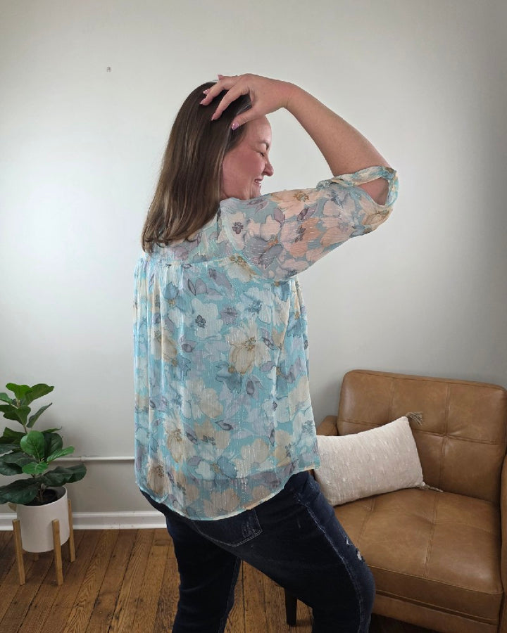 A woman with long hair stands smiling in a room, wearing the Kaley Floral 3/4 Sleeve Blouse with Sheer Overlay and dark jeans, both from Cosmos and Clover Boutique. The wooden-floored space features a tan couch, potted plant, and a plain light wall.