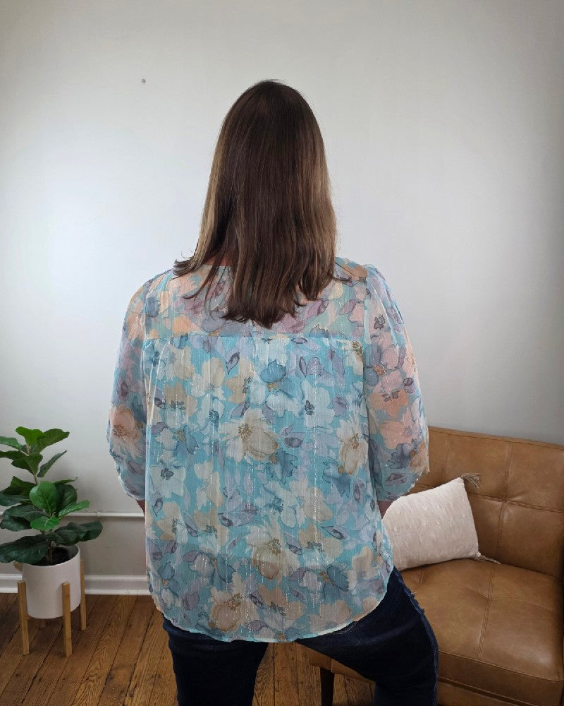 A woman with long hair stands smiling in a room, wearing the Kaley Floral 3/4 Sleeve Blouse with Sheer Overlay and dark jeans, both from Cosmos and Clover Boutique. The wooden-floored space features a tan couch, potted plant, and a plain light wall.