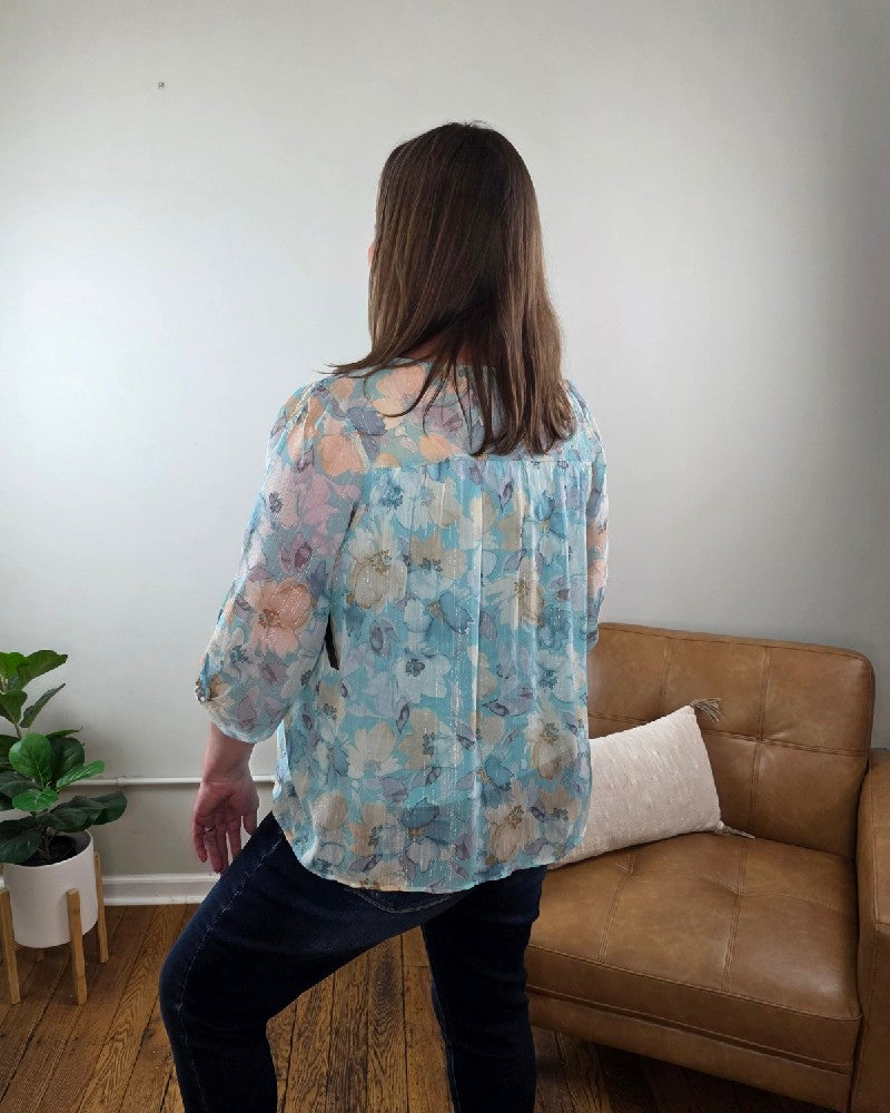 A person with long brown hair, wearing the Kaley Floral 3/4 Sleeve Blouse with Sheer Overlay and dark jeans, stands facing away in a room with a tan leather chair, pillow, plant, and wood floor.