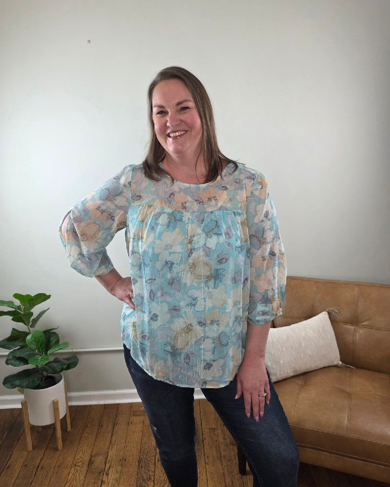 A woman with long hair stands smiling in a room, wearing the Kaley Floral 3/4 Sleeve Blouse with Sheer Overlay and dark jeans, both from Cosmos and Clover Boutique. The wooden-floored space features a tan couch, potted plant, and a plain light wall.