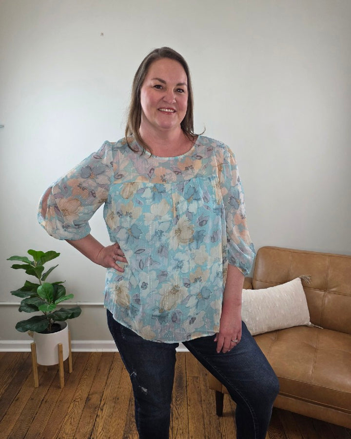 A woman with long hair stands smiling in a room, wearing the Kaley Floral 3/4 Sleeve Blouse with Sheer Overlay and dark jeans, both from Cosmos and Clover Boutique. The wooden-floored space features a tan couch, potted plant, and a plain light wall.