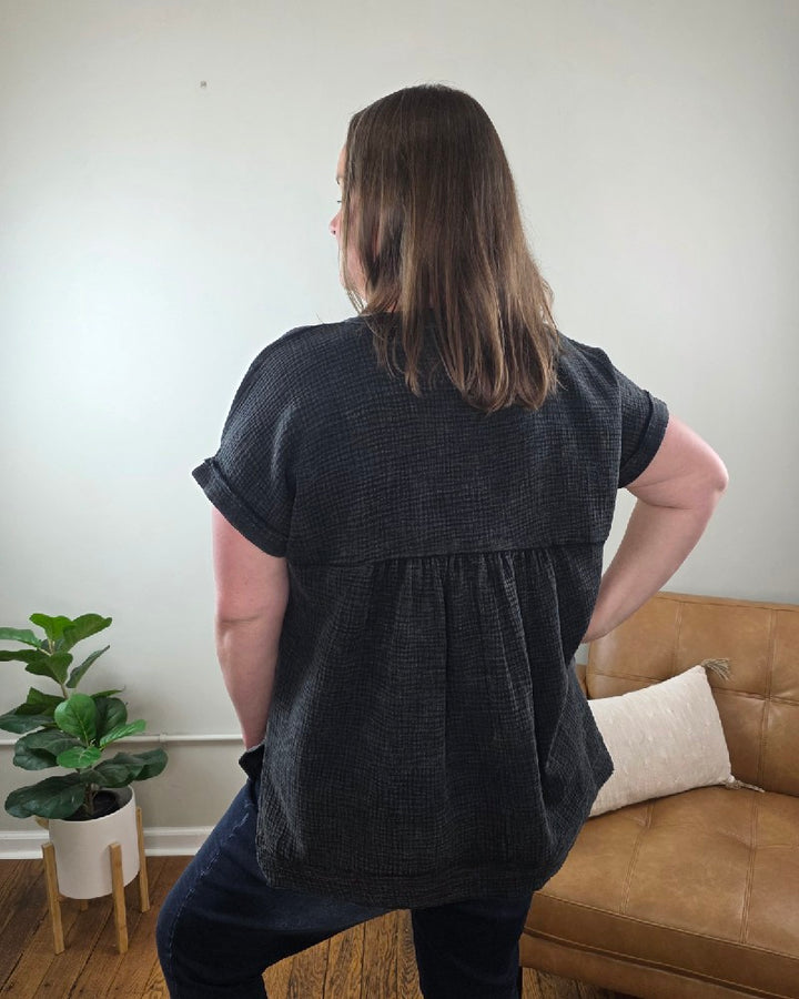 A woman with straight, shoulder-length hair wears the ZENANA Kasey Short Sleeve Washed Henley Shirt and jeans, smiling in a living room featuring a plant, brown leather couch, and white pillow in the background.