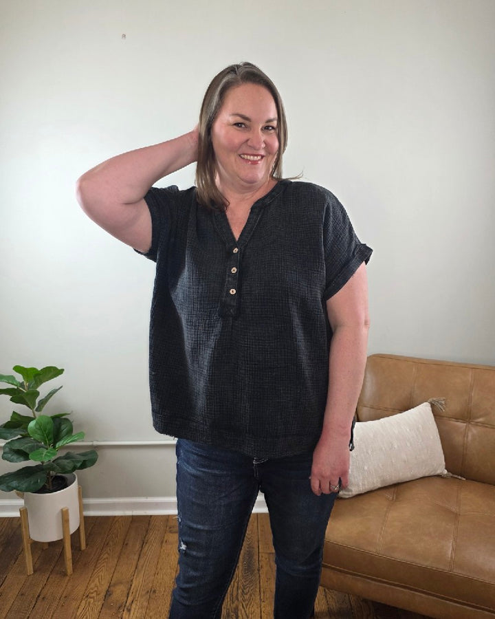 A woman with straight, shoulder-length hair wears the ZENANA Kasey Short Sleeve Washed Henley Shirt and jeans, available at Cosmos and Clover Boutique, smiling in a living room featuring a plant, brown leather couch, and white pillow in the background.