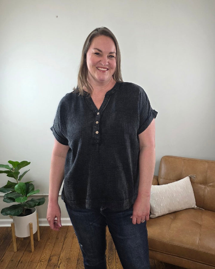 A woman with straight, shoulder-length hair wears the ZENANA Kasey Short Sleeve Washed Henley Shirt and jeans, smiling in a living room featuring a plant, brown leather couch, and white pillow in the background.