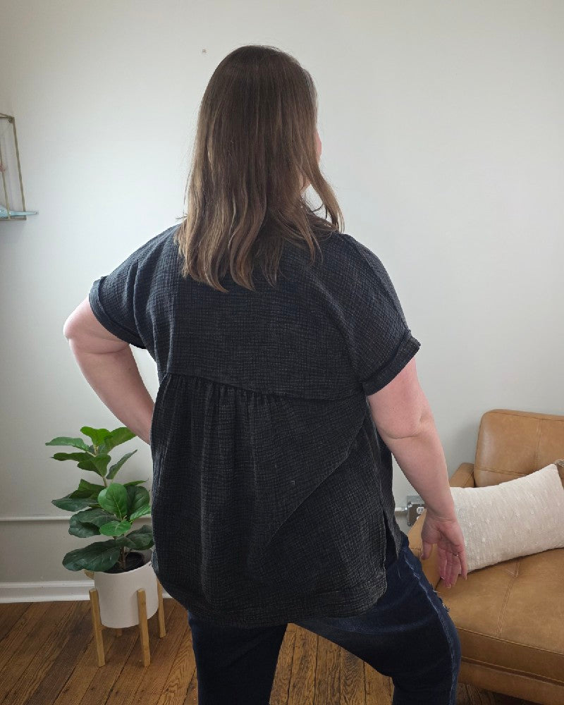 A person with shoulder-length brown hair stands indoors, one hand on their hip, wearing the ZENANA Kasey Short Sleeve Washed Henley Shirt and jeans. A brown chair and a green potted plant in the background enhance the relaxed vibe.