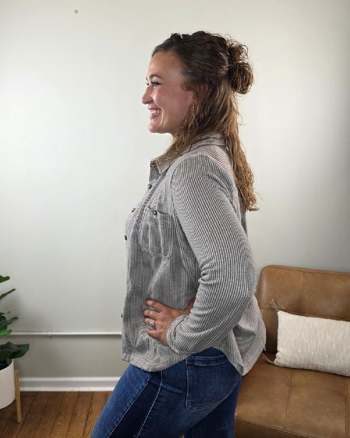 A woman stands in profile with a smile, her hand resting on her hip. She wears the blu pepper Kate Button Down Shacket in gray, paired with blue jeans—its relaxed fit makes it an ideal layering piece. In the background, a tan couch with a cushion and a green plant in a white pot sit against a light-colored wall.