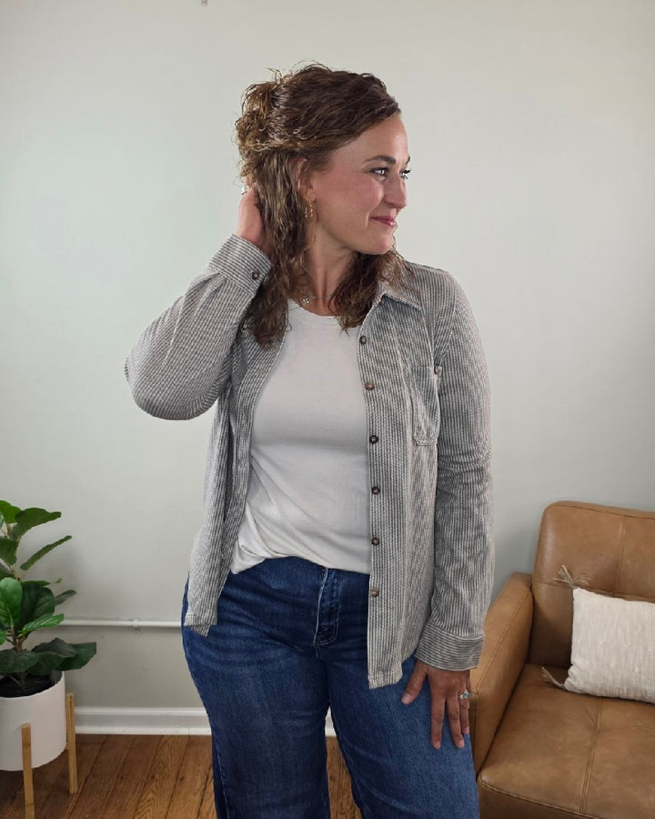 A woman with curly hair stands indoors, smiling and touching her hair while wearing the blu pepper Kate Button Down Shacket—a relaxed fit, striped layering piece—over a white top and blue jeans. A plant and a tan couch accent the background.