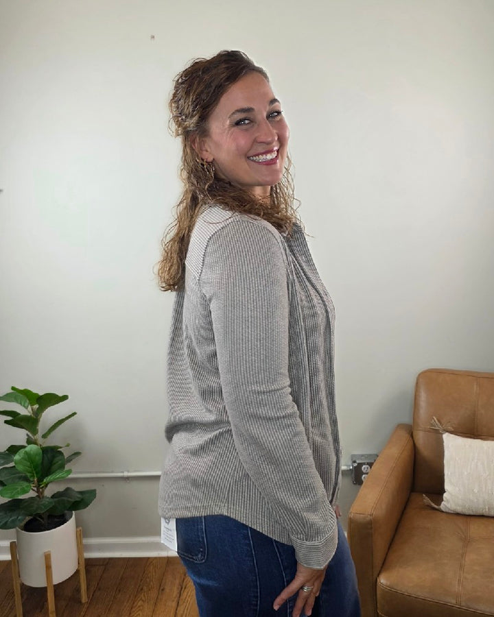 A woman with curly hair stands smiling in a living room styled with a tan chair, white pillow, and potted plant, wearing the blu pepper Kate Button Down Shacket paired with jeans.