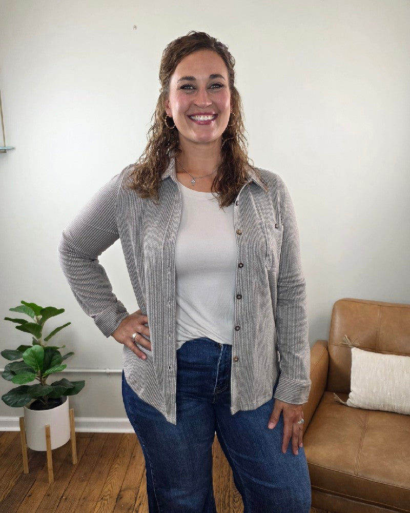 A woman with curly hair smiles at the camera while standing indoors. She wears the Kate Button Down Shacket by blu pepper—a relaxed fit, striped piece—layered over a white tee and blue jeans. In the background, a brown couch with pillows and a potted plant can be seen.