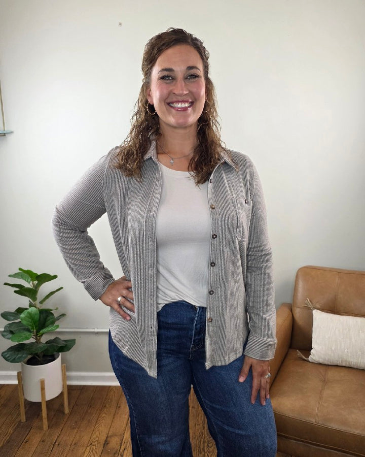 A woman with curly hair smiles at the camera while standing indoors. She wears the Kate Button Down Shacket by blu pepper—a relaxed fit, striped piece—layered over a white tee and blue jeans. In the background, a brown couch with pillows and a potted plant can be seen.
