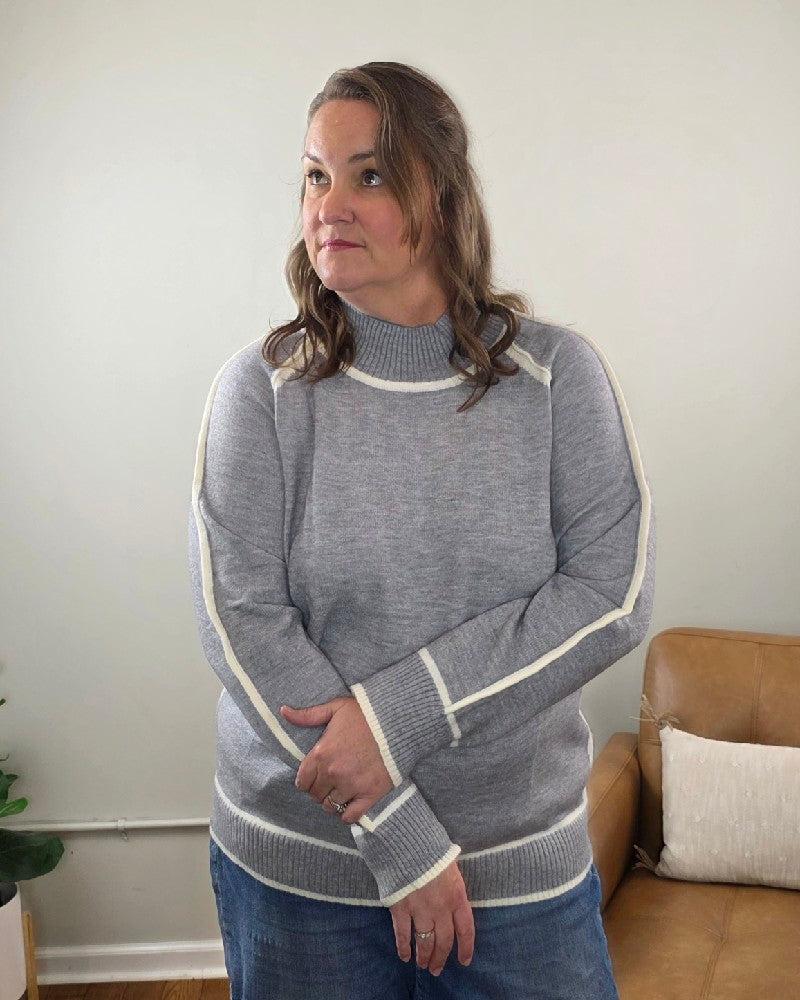A woman wears a grey Kathy Sweater by Lime N Chili, Inc, featuring a mock neck and white striped trim along the sleeves and cuffs.