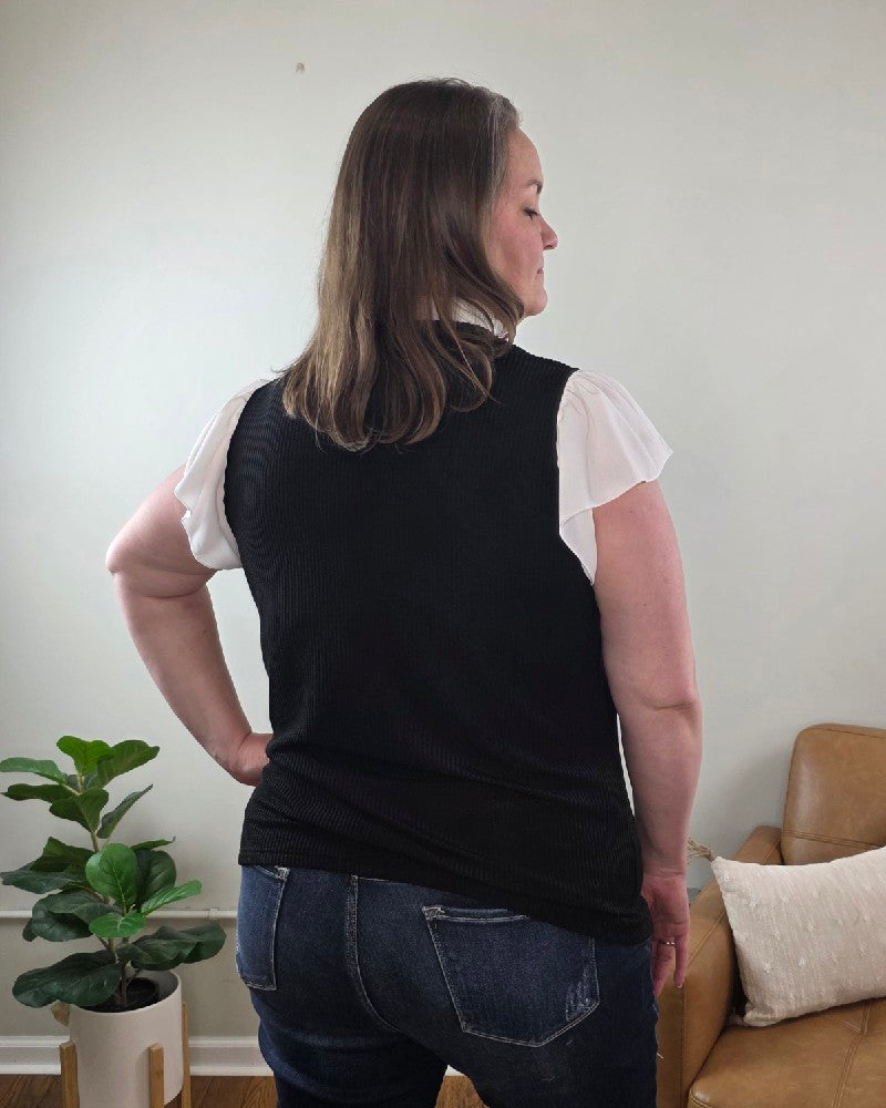 A woman with long hair smiles at the camera indoors, wearing the Katie Ruffle Sleeve Ribbed Blouse and blue jeans, standing near a light brown sofa with a white pillow.