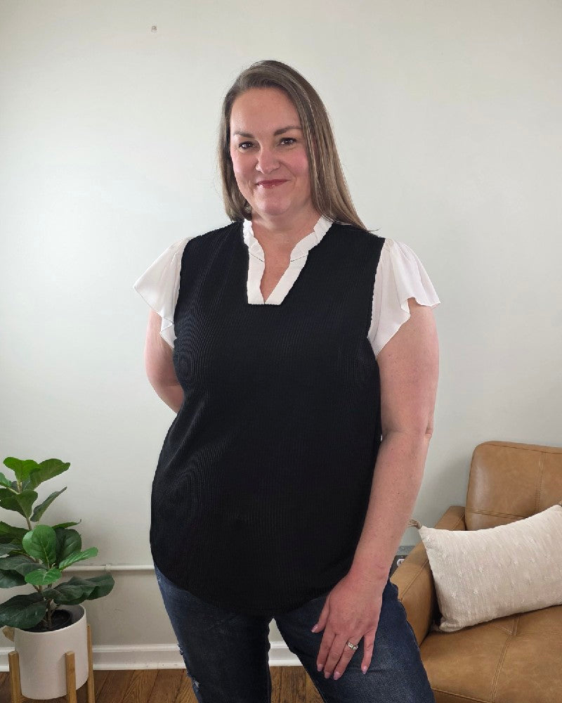 A woman with long hair smiles at the camera indoors, wearing the Katie Ruffle Sleeve Ribbed Blouse and blue jeans, standing near a light brown sofa with a white pillow.
