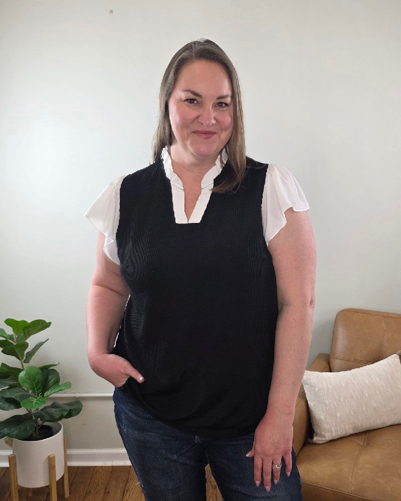 A woman with long hair smiles at the camera indoors, wearing the Katie Ruffle Sleeve Ribbed Blouse and blue jeans, standing near a light brown sofa with a white pillow.