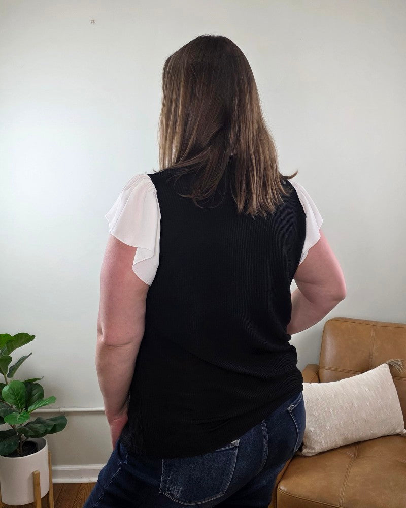 A woman with brown hair, seen from behind, stands indoors wearing the Katie Ruffle Sleeve Ribbed Blouse in black and white with blue jeans, beside a tan couch with a pillow and a potted plant.