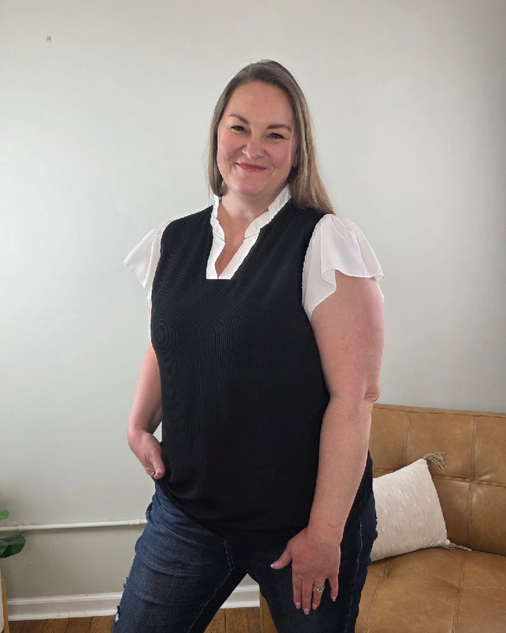 A woman with long hair smiles at the camera indoors, wearing the Katie Ruffle Sleeve Ribbed Blouse and blue jeans, standing near a light brown sofa with a white pillow.