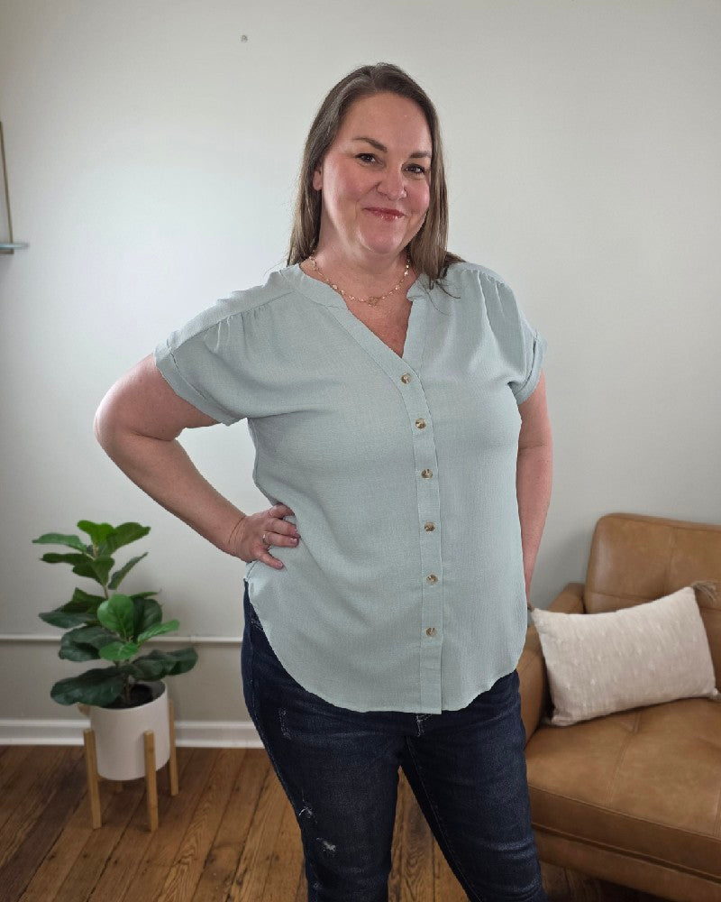 A woman with long brown hair stands indoors, smiling with one hand on her hip. She wears the Kayla Rolled Sleeve Button Down Top and dark jeans, with a potted plant, tan couch, and light wall in the background.