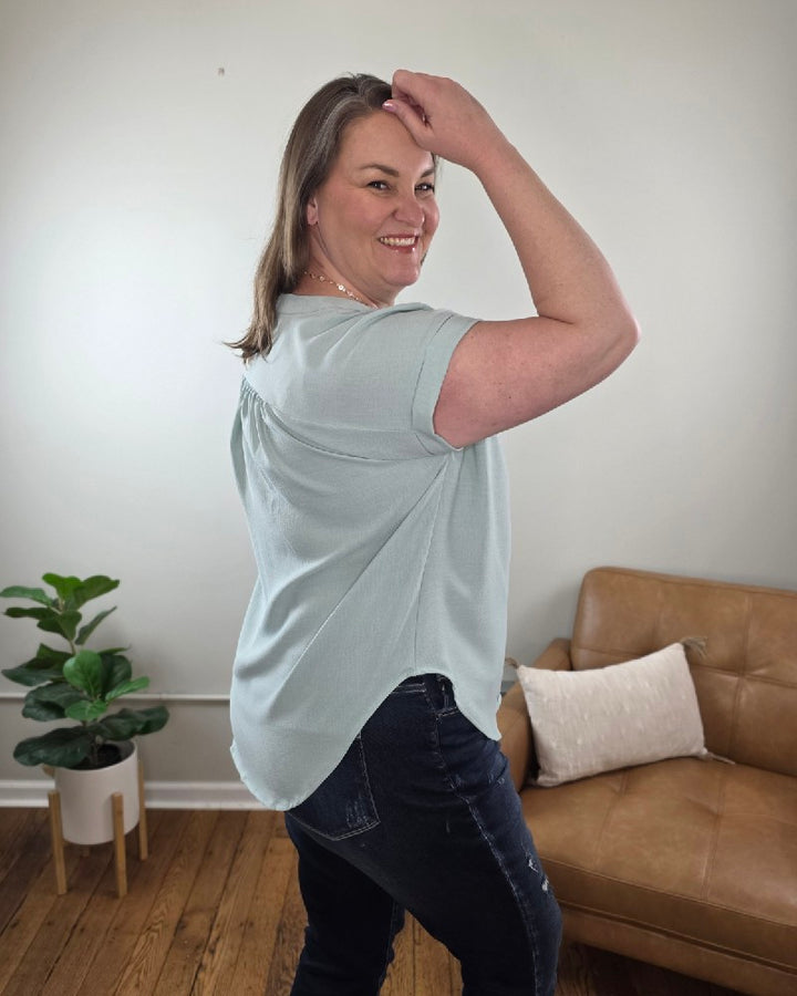 A woman with shoulder-length brown hair stands indoors in the Kayla Rolled Sleeve Button Down Top from Cosmos and Clover Boutique, light blue, paired with dark jeans. A brown sofa, white pillow, and potted plant emphasize this versatile blouse for any casual setting.