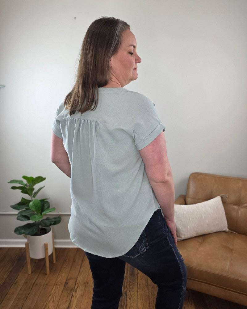 A woman with shoulder-length brown hair stands indoors in the Kayla Rolled Sleeve Button Down Top from Cosmos and Clover Boutique, light blue, paired with dark jeans. A brown sofa, white pillow, and potted plant emphasize this versatile blouse for any casual setting.