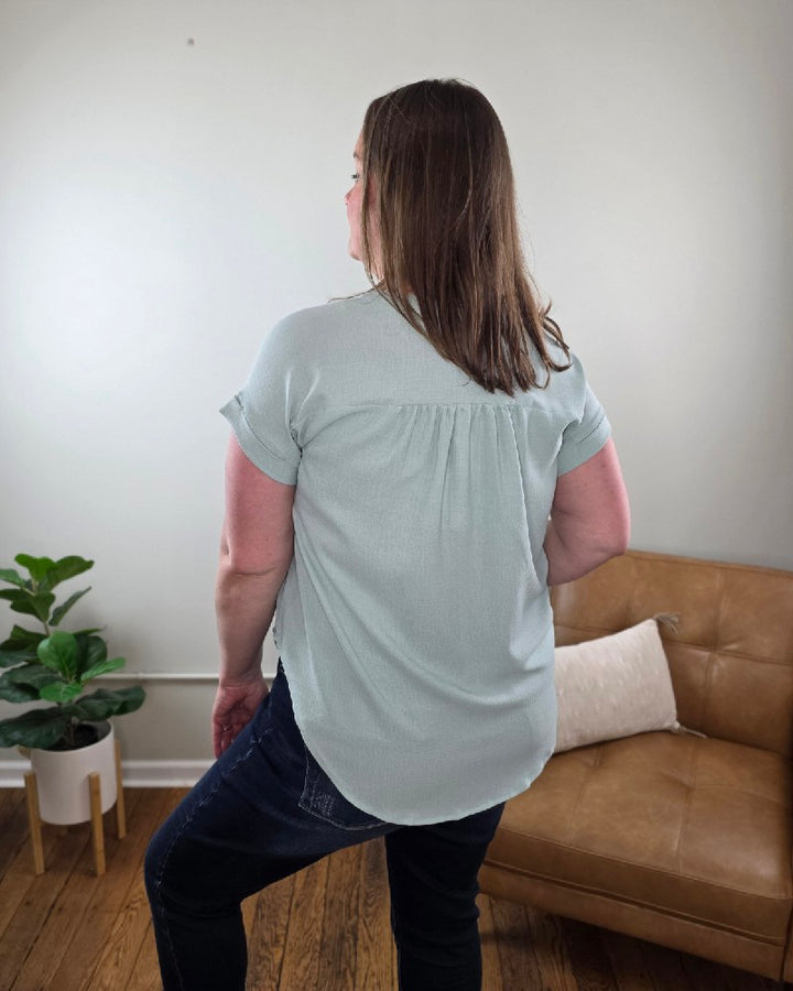 A woman with shoulder-length brown hair stands indoors in the Kayla Rolled Sleeve Button Down Top from Cosmos and Clover Boutique, light blue, paired with dark jeans. A brown sofa, white pillow, and potted plant emphasize this versatile blouse for any casual setting.