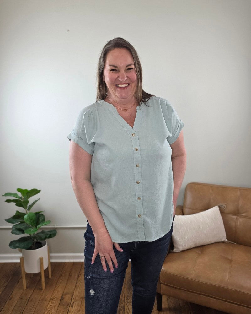 A woman with shoulder-length brown hair stands indoors in the Kayla Rolled Sleeve Button Down Top from Cosmos and Clover Boutique, light blue, paired with dark jeans. A brown sofa, white pillow, and potted plant emphasize this versatile blouse for any casual setting.