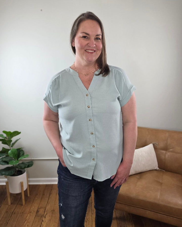 A woman with shoulder-length brown hair stands indoors in the Kayla Rolled Sleeve Button Down Top from Cosmos and Clover Boutique, light blue, paired with dark jeans. A brown sofa, white pillow, and potted plant emphasize this versatile blouse for any casual setting.