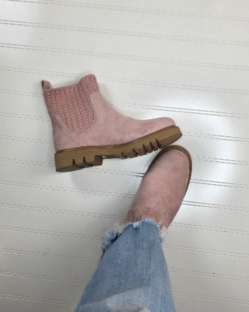 A person wearing light blue jeans and pink Keep Me Warm Boot by Corkys with thick, textured soles stands on a white, paneled floor. One boot is on the floor, while the other is slightly lifted, showcasing the cozy cable knit upper.