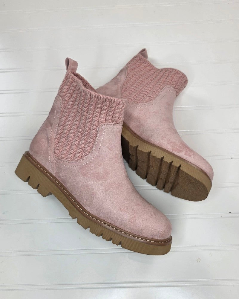 A pair of Corkys Keep Me Warm Boots in light pink, featuring chunky tan rubber soles, knitted elastic side panels, and a cozy cable knit upper, are displayed on a white wooden surface.