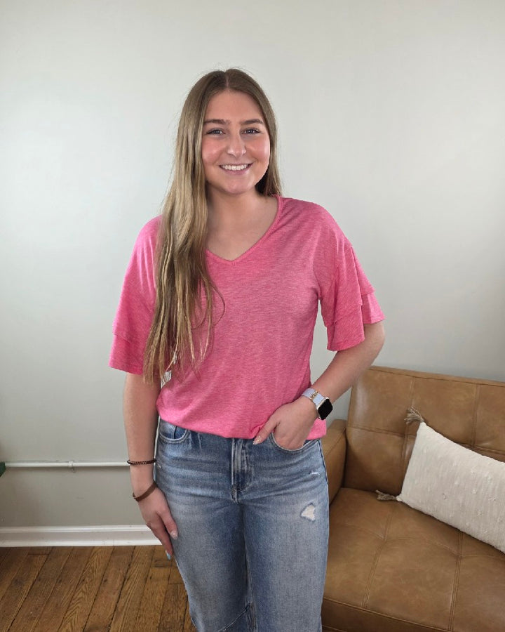 A young woman with long brown hair stands indoors, smiling. She is wearing the Lovesoft Kia V-Neck Ruffle Sleeve Top, light blue jeans, and a smart watch in front of a tan couch against a light-colored wall.