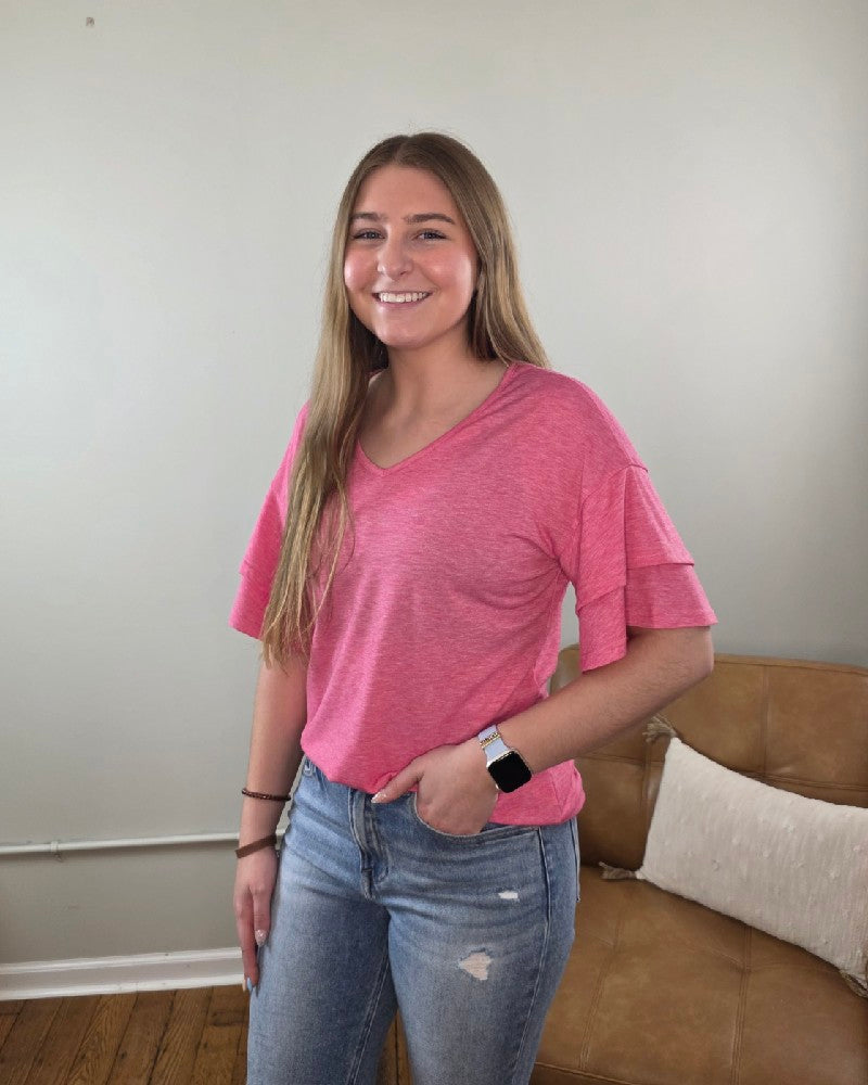 A young woman with long hair, wearing the Lovesoft Kia V-Neck Ruffle Sleeve Top and ripped blue jeans, stands smiling in a living room with a beige couch and light-colored wall in the background.